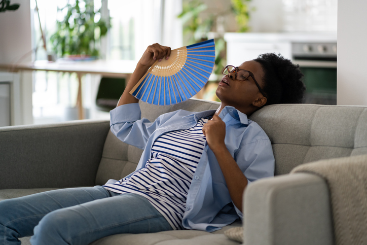 Tired exhausted woman sits on sofa at home using paper fans and closing eyes suffering from stuffiness. Tormented black girl housewife is resting after hot walk in summer weather